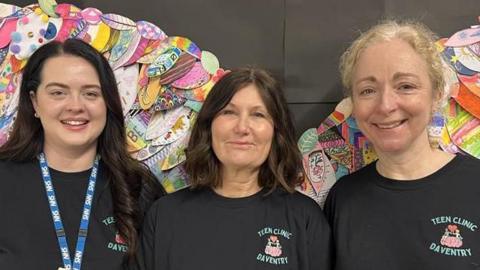 Three women stood side-by-side in black T-shirts. All are looking at the camera and smiling. Behind them is a colourful display.