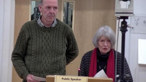 A tall older man and a short older woman stand up at a council meeting, addressing the room. The woman is speaking as the man looks around the room, looking serious. The podium they are standing at reads "public speaker" and has a small, sleek microphone attached to it.