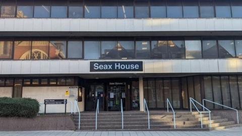 The entrance to Essex Coroner's Court at Seax House - a photo taken from the other side of the road. There are steps and white-painted railings leading up to the court entrance, and the sign on the 1960s multi-storey building reads SEAX HOUSE.