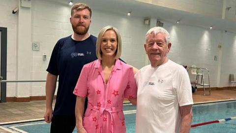 Kay Crewdson is standing at the side of an indoor swimming pool next to Ken and his grandson Sam. Both of the men are wearing T-shirts and their swimwear