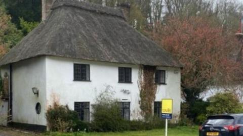 A two-storey white thatched cottage surrounded by trees. There are three windows on each floor.