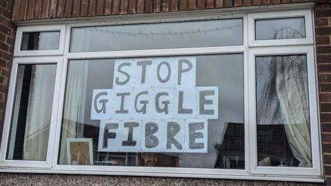 A house window with a sign that reads 'Stop Giggle Fibre' and a man's smiling face beneath