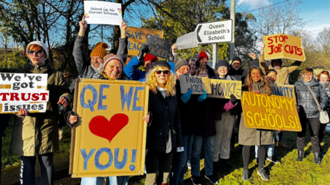 A group of people wearing hats and coats stand on the grass outside QE School holding signs saying "QE we love you" and "Stop job cuts".