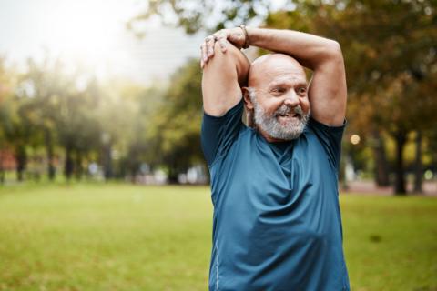 Man stretching with arms above head in a park with green grass. The man has a grey beard, is bald and wears a blue t-shirt. He is smiling. 