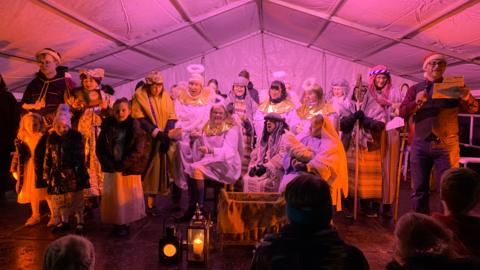 A Christmas nativity scene cast gathered on a stage under a marquee. There are children and adult actors. Some women are dressed as angels and some men as wisemen in the centre of the group, with a manger in front of them. 