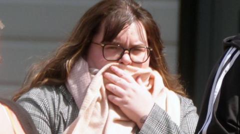 A woman with long brown hair and glasses covers her face with a scarf outside of court