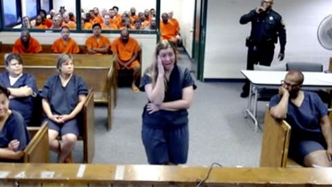 A woman in a blue prison jump suit wraps one arm across her stomach and one hand to her face. There are other female prisoners sitting on wood benches in similar jumpsuits. There are a group of men in orange jumpsuits on wood benches in the background of the image.