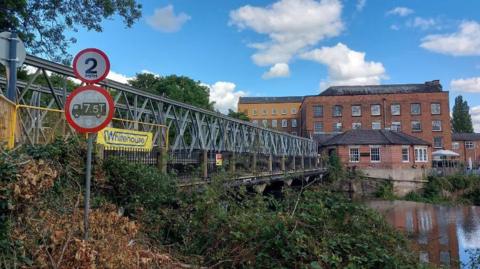 An iron bridge which stretches over a body of water. Several red-brick buildings can be seen in the background on the far side of the water.