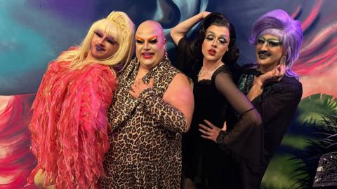 Four drag artists posing for the camera in front of a mural. From left to right: One is wearing pink feathers and a blonde wig; one in leopard print; a dark haired drag artist in a black laced dress and pearls; and on the end in black, is one with a purple wig and moustache.