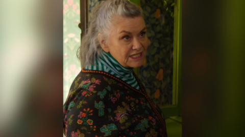 A woman in a floral patterned cardigan and striped top looks at someone off camera with an annoyed expression