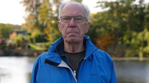 A man wearing a blue coat looks into the camera with a neutral expression on his face. He is standing next to a river.