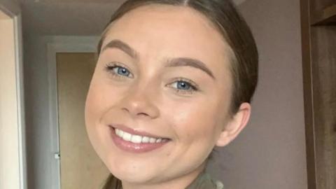 Jaysley Beck smiling into camera, wearing military unitform. She has dark hair and blue eyes. A light coloured door in an inside room is behind her.