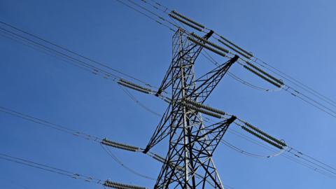 An electricity tower, or pylon, pictured looking up it from the ground and against a clear blue sky. The tower is a lattice-work of metal with "arms" sticking from the side attached to cables.