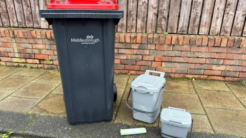 A black wheelie bin with a red lid and the words 'Middlesbrough moving forwards' in white text on the front on a pavement with a wall and fence behind it. To its right are two small grey waste caddies and a roll of bin bags.
