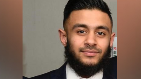 A man wearing a suit and pink tie looks at the camera. He has black hair and a thick beard.