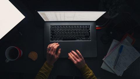 Aerial view of hands typing on a laptop, with a cup of tea, a biscuit, and notepads filled with notes on a dark desk.