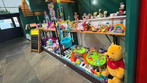 An array of children's toys from a Winnie the Pooh cuddly toy to a toy phone are displayed on three shelves. The wall behind the display is teal and has vertical white boards along the bottom half.