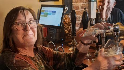 Sally Ward standing behind the bar of a pub pulling a pint. She is looking directly at the camera and is wearing a green, orange and brown patterned long sleeved T-shirt. She has a pair of glasses on her eyes.