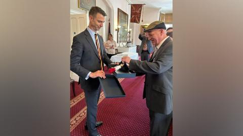 Major General Jon Swift, dressed in a dark grey suit, presents Fusilier Steve Close, also wearing a grey suit, with a presentation box containing his beret and hackle, made up of red and white feathers. Several people are smiling in the background