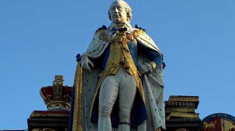 Statue of The King, in Garter robes backed by various insignia, including, to his right, the crown on a cushion, Royal standard and Union flag. 