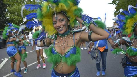 A dancer wearing a yellow and green feathered headdress and a blue bikini top with green feathers on it smiles into the camera as she takes part in a parade in the Caribbean Carnival