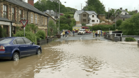 Pwll o ddwr brown yn sgwar Tal-y-bont, Ceredigion - cerbydau a swyddogion y gwasanaeth tan yn y cefndir. Pobl yn edrych ar y pwll o ddwr.