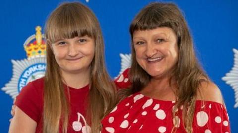 Lulu and her mother Tracey stand in front of a blue backdrop with the North Yorkshire Police logo on it. Lulu has long brown hair and a fringe, and wears a red t-shirt with a white cherry logo on the front. Ms Frost has long brown, wavy hair and a fringe and wears a red off the shoulder top with white spots.