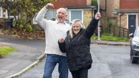 Man in white jumper and woman in dark coat both with hands in the air in celebration stading in a cul de sac with houses behind them