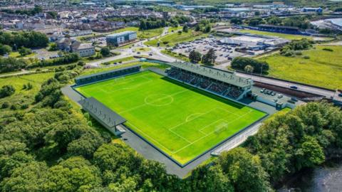A stadium pitch, surrounded by trees and a large car park with houses in the distance.