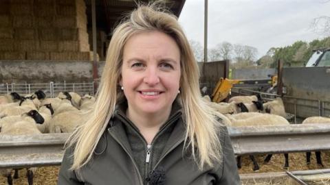 Isobel smiles for the camera, her sheep in a pen behind her. She has long blonde hair and wears a browny green jacket and fleece.