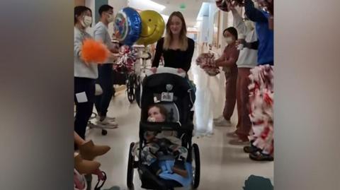 Boy wheeled in pram through corrider by mother surrounded by clapping nursing staff
