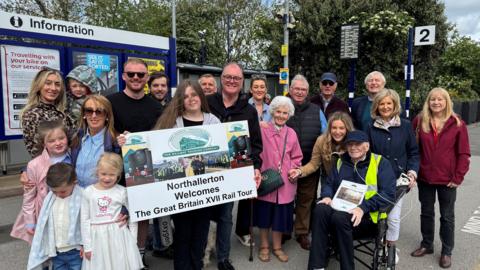 An elderly man in a wheelchair with his family. About 20 people including children. They are stood on a railway platform holding a sign that reads 'Northallerton Welcomes The Great Britain XVII Rail Tour'.