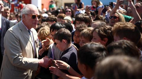 King Charles III in sunglasses, wearing a beige suit with military medals. He is shaking hands with a group of children. Adults smile and take photographs in the background.