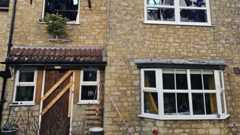 A stone built house with a bay window on the right hand of the image and a small porch with a front door flanked by two small windows on the left hand of the image. All the glass has gone from the windows due to fire damage and their are wooden slats covering the front door of the house