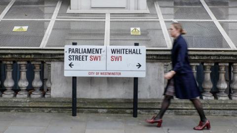 A pedestrian passes a street sign display the start of Parliament Street, left, and Whitehall, right, in the Westminster district of London, UK.