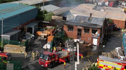 A view of a fire-damaged commercial premises, taken from a high vantage point. Several fire engines and firefighters are at the scene in front of the building.