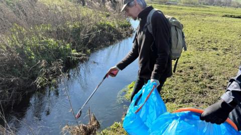 A volunteer from Wear Rivers Trust stands by a stream and is picking up waste with a litter-picker. They are holding a bright blue bin bag. The volunteer has short grey hair and is wearing all black outdoor clothing, a grey cap and a grey rucksack.