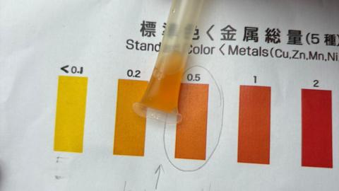 A test sheet, showing different colours on a chart, with writing above and a water sample in orange, matching one of the colours on the chart. There is writing on the paper in English and another language. 