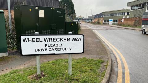 A road sign which has been covered up with the words "Wheel Wrecker Way, Please! Drive Carefully". A road with potholes is in the background with an industrial estate beyond. The first section of the road is maintained with no potholes.