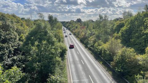 An aerial view of the A361 Frome Bypass on a cloudy day. The road has a northbound and southbound lane and far cars can be seen driving along the long stretch of road. The road is surrounded by tall green trees.