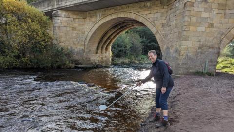 A woman wearing trousers and a blue top is taking a water sample from the River Coquet which is wide and quite fast flowing. She is just in front of a old bridge 