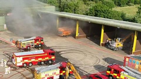 A number of fire engines on a concrete surface with thick smoke hanging in the air.