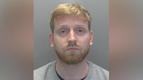 A police mugshot of Jakob Ireson. He has short ginger hair and beard, and is standing in front of a grey wall. He is wearing a grey jumper with a black T-shirt underneath.