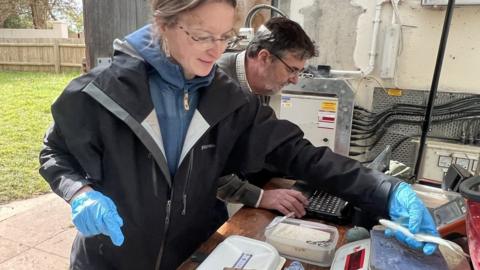 Fisheries ecologist Sophie Elliott placing a juvenile salmon on a scale with fisheries scientist Bill Beaumont in the background entering data into a laptop
