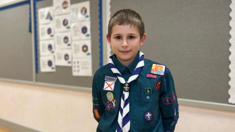 Charlie looks at the camera, he is wearing his scouts uniform which is a dark green shirt with a blue and white neckerchief. He has several bright badges sown onto his uniform. Charlie has dark, brown eyes and short light brown hair.