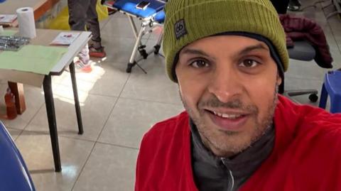 Gautam Sharma is standing in a dentistry clinic. He is wearing a grey zip up jacket with a red jacket over the top. He is wearing an olive green beanie hat and is looking directly at the camera, taking a selfie. 