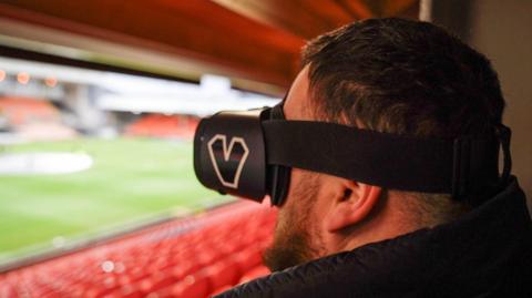 Jon Attenborough, a man with short dark hair and a beard, looks over an empty football stadium while wearing a headset that covers his eyes