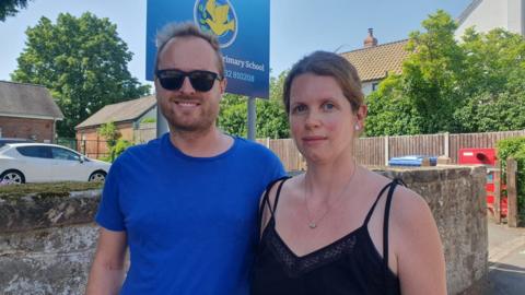 A man in a blue t shirt and a woman in a black strapped vest standing side by side with a wall and school sign in the background