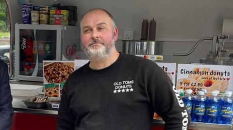 A man with a grey beard in a black top at a food van. Cans are enclosed behind him and a sign on the wall lists hot drinks.
