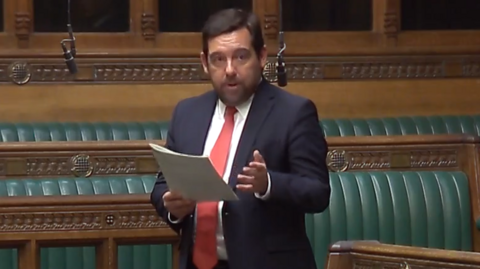 David Baines is standing in parliament. He has short brown hair, and is wearing a red tie, white shirt, and navy jacket. He is speaking, and reading from a sheet of paper.